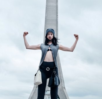 Person iført en grå vest, sort crop top og leggings, der spænder armene foran et højt beton tårn under en overskyet himmel.