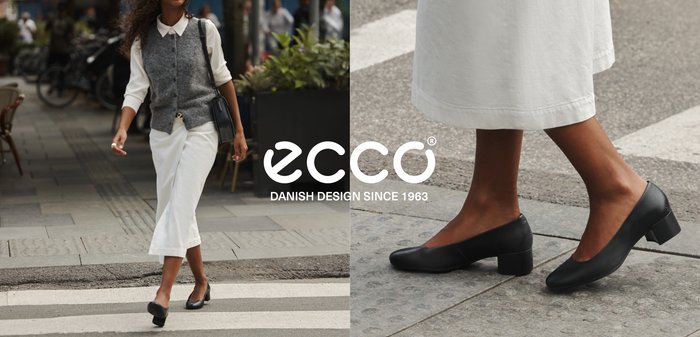 Femme traversant un passage piéton portant des talons noirs, un pantalon blanc à jambes larges, une chemise blanche, un gilet gris, et portant un sac à main noir.