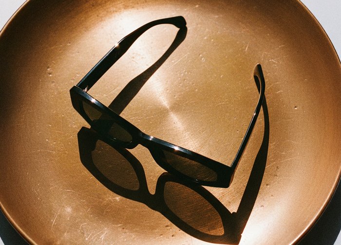 Lunettes de soleil noires avec une monture rectangulaire reposant sur une plaque circulaire en bronze, projetant des ombres marquées sur la surface.