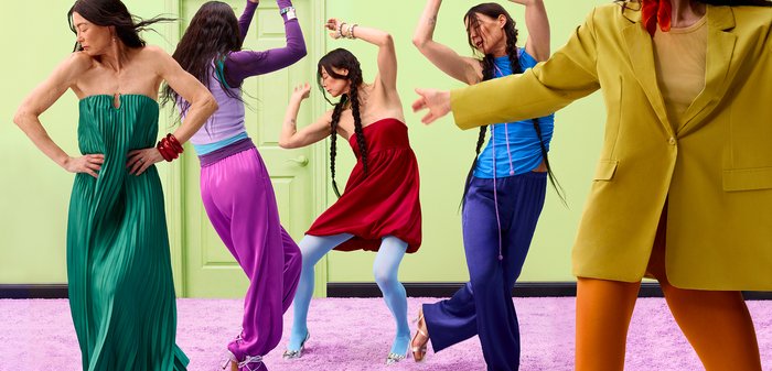 Cinq personnes en tenues colorées dansant avec énergie dans une pièce verte avec un tapis rose.