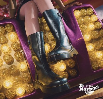 Botas de cuero negro hasta la rodilla con un acabado brillante, costuras amarillas y suela gruesa, exhibidas contra un fondo de luces circulares.