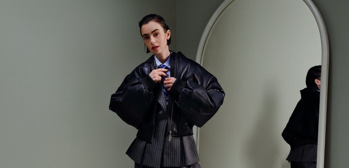 Vrouw die een zwarte oversized leren jas draagt over een krijtstreeppak, terwijl ze de jas aanpast en naast een grote boogvormige spiegel staat.