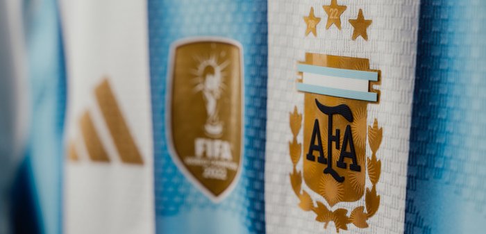 Maillot de football argentin avec emblème de l'AFA, badge doré des champions de la Coupe du Monde FIFA 2022, logo Adidas, et rayures bleu et blanc.