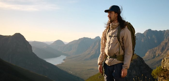 Man wearing a cap and beige jacket with backpack, standing outdoors in mountainous landscape during clear daylight.