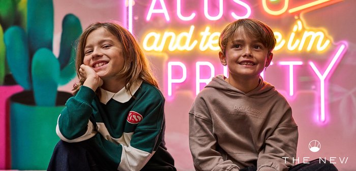 Deux enfants souriants assis à l'intérieur avec un panneau néon coloré et une grande plante cactus en arrière-plan.