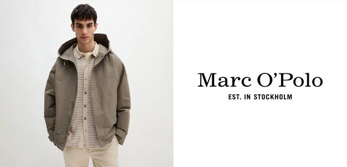 Młody mężczyzna w beżowych spodniach, jasnym swetrze z dzianiny i taupe'owej kurtce z kapturem, stojący na tle jednolitego białego tła.