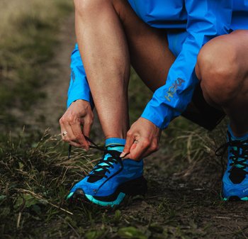 Des chaussures de course Hoka en bleu avec des accents noirs et des semelles turquoise. La personne lace les chaussures tout en portant un haut à manches longues bleu.