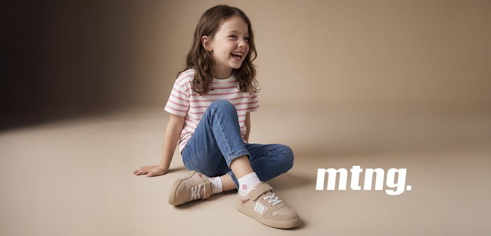 Niña sonriente con cabello rizado y castaño sentada con las piernas cruzadas, viste una camiseta de rayas rosa, jeans azules, calcetines con estampado de flores y zapatillas beige con logo "m".