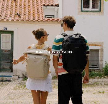 Dva lidé v slunečních brýlích s velkými batohy kráčí po dlážděné ulici vedle bílých budov s taškovými střechami.