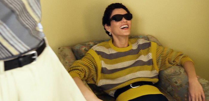 Person iført sorte solbriller og en sweater med gule striber smiler, mens de læner sig tilbage på en mønstret sofa mod en beige væg.