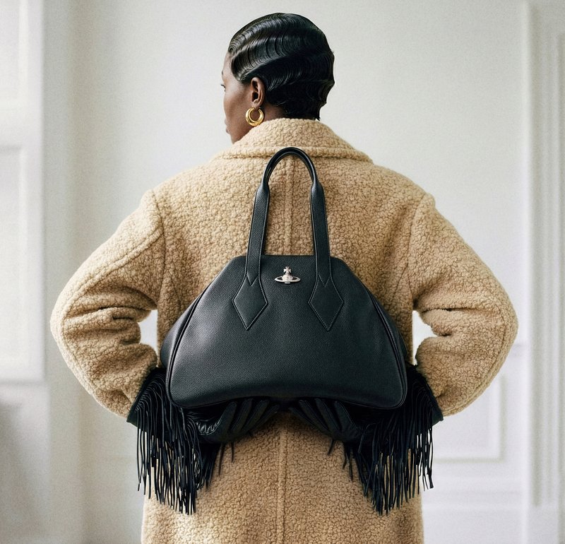 Person iført en beige tekstureret frakke og guldringe øreringe holder en sort læderhåndtaske med frynser bag ryggen.