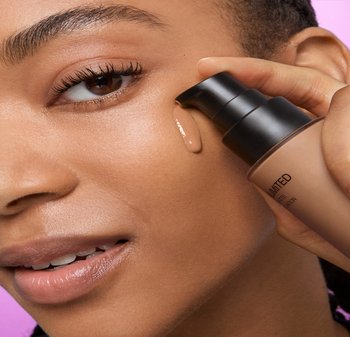 Liquid foundation in a pump bottle with a matte finish, held against a model's skin. Brown tone, smooth texture, highlighted by a pink backdrop.