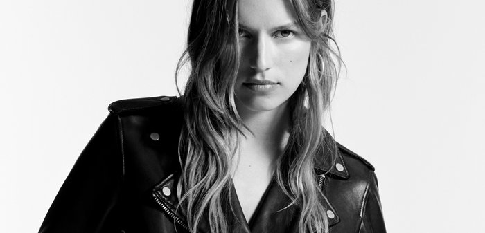 Jeune femme aux longs cheveux portant une veste de motard en cuir noir avec des poches zippées, regardant droit devant avec les mains dans les poches.