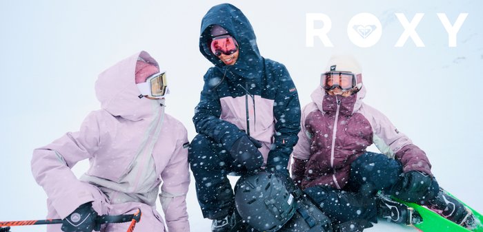 Trois personnes en vestes de ski colorées : l'une en rose clair, l'autre en marine avec des accents rose clair, et une troisième en bordeaux profond, portant des lunettes de ski.
