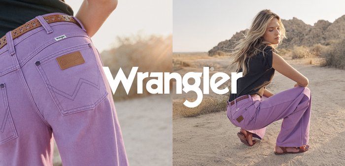 Kvinne i svart topp og lavendelfargede jeans sitter på huk på en ørkensti med steinete åser i bakgrunnen, Wrangler-logoen plassert midt i bildet.