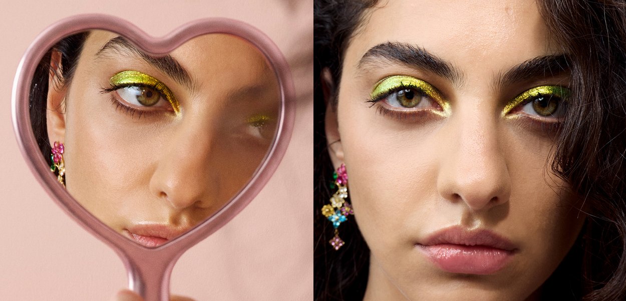 Femme portant un fard à paupières jaune-vert vif, des boucles d'oreilles colorées, et des cheveux bouclés foncés, montrée dans un miroir en forme de cœur avec un gros plan de face.