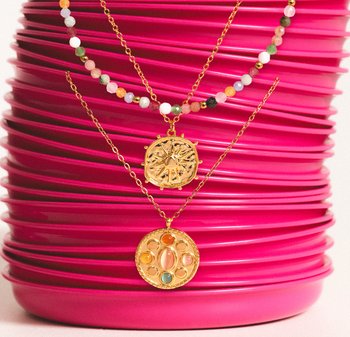 Deux colliers en or présentent des pendentifs : l'un circulaire avec des pierres colorées, l'autre en forme de boussole. Les deux pendentifs pendent de chaînes fines en or.
