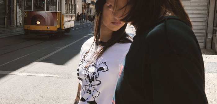 Two people stand near a street with an approaching yellow tram; one wears a white shirt with a cartoon bunny design.