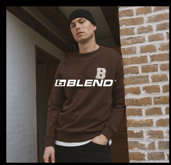 Bruine sweat met een wit "B"-embleem, ronde hals en geribbelde boorden, gedragen met een wit hemd eronder. Structuur van een bakstenen muur op de achtergrond.