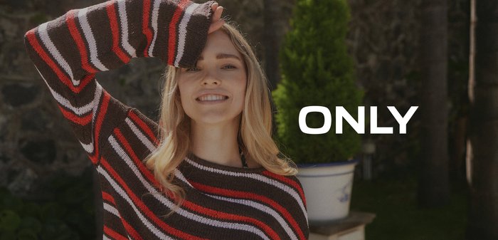 Tânără zâmbitoare în aer liber, purtând un pulover cu dungi maro, roșu, alb și roz, cu un braț ridicat deasupra capului.