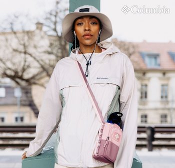 Leichte, zweifarbige Columbia-Jacke in Beige und Grün mit verstellbarem Kapuzen. Rosa Umhängetasche, die eine blaue Wasserflasche enthält.