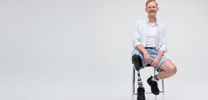 Chemise rayée bleue clair, débardeur blanc, shorts en denim. Chaussures de sport noires et chaussettes rayées. Inclut une prothèse de jambe sur une chaise en métal.