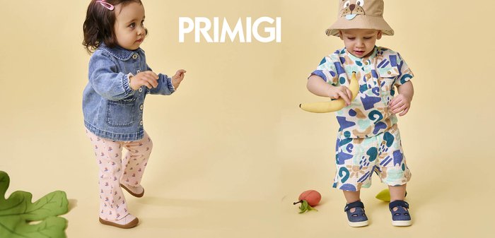 Two toddlers on beige background; girl in denim jacket and pink pants faces boy in patterned outfit and animal hat holding a banana toy.