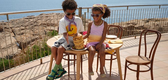 Deux enfants portant des lunettes de soleil sont assis à une table au bord de la mer avec une grande pile de pancakes entre eux sur une assiette.