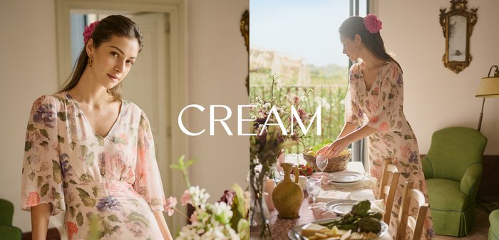 Frau in einem cremenen, geblümten Kleid steht neben einem Tisch mit einem Korb Orangen und Vasen mit Blumen in einem sanft beleuchteten Raum.