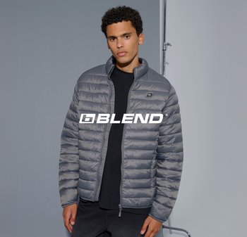 Giovane uomo in giacca grigia trapuntata e maglietta nera in piedi contro uno sfondo grigio con il logo "BLEND" sul petto.