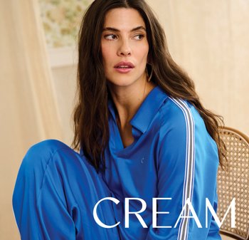 Woman with long dark hair wearing a blue tracksuit with white stripes, seated on a woven chair, looking to her right.