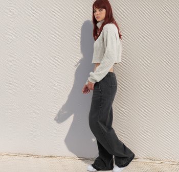 Geknipte, lichtgrijze kabelgebreide trui met hoge hals; gecombineerd met losse zwarte jeans en witte sneakers. Gestructureerde achtergrond.