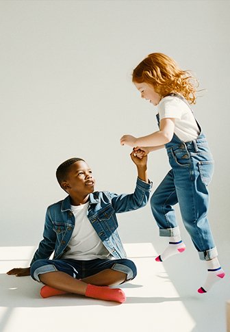 Duas crianças brincam juntas. Uma veste uma jaqueta de ganga e calções, enquanto a outra está com um macacão de ganga e uma t-shirt branca, meias com detalhes em rosa.