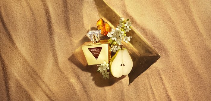 Butelka perfumu marki Guess, plaster gruszki, białe kwiaty, bursztynowy kryształ na złotej trójkątnej tacy na tle fakturowanego piasku.