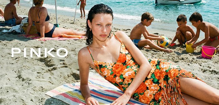 Giovane donna in un vestito arancione floreale sdraiata su un asciugamano da spiaggia con bambini che giocano con una palla vicino al mare sullo sfondo.