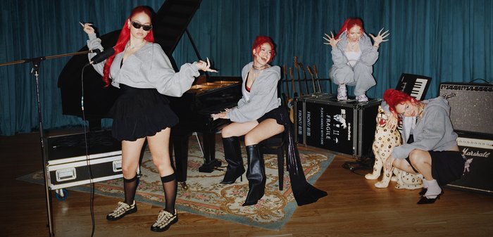 Vier Frauen mit roten Haaren in grauen Kapuzenpullovern und schwarzen Röcken posieren mit Klavier, Gitarren und Verstärkern in einem Musikstudio mit Holzboden.