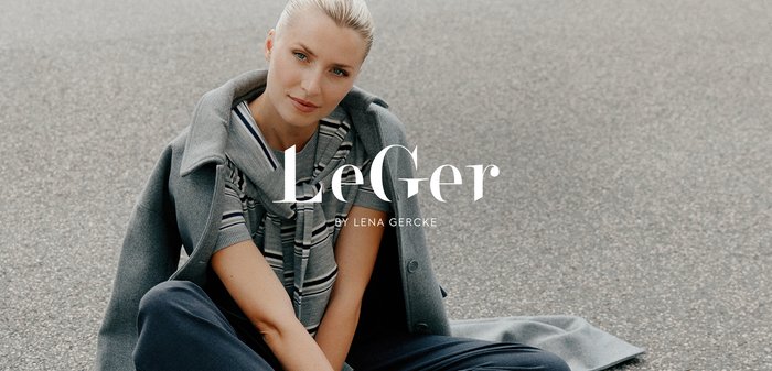 Cappotto oversize grigio chiaro su una maglia a righe, abbinato a pantaloni scuri. Il soggetto è seduto, mostrando un anello sulla mano.