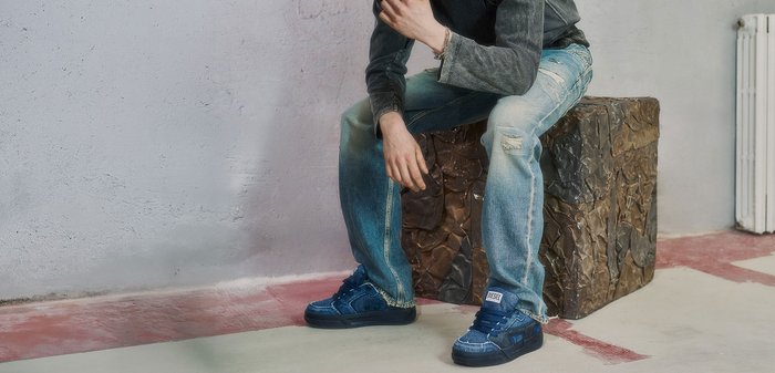 Person mit blondem Haar trägt ausgewaschene Jeans, dunkle Jacke und blaue Turnschuhe und sitzt auf einem strukturierten braunen Würfel drinnen.