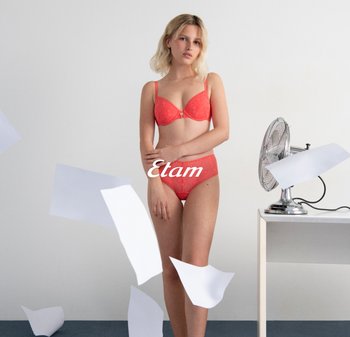 Rote Spitzen-BH und passender Slip, präsentiert an einem stehenden Modell. Im Hintergrund befinden sich verstreute Papiere und ein silberner Ventilator auf einem weißen Tisch.