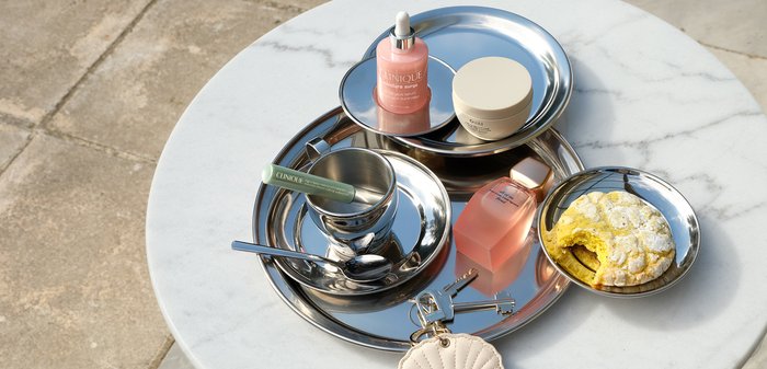 Des plateaux ronds en argent sur une table en marbre blanc contiennent des produits de soin de la peau, un baume à lèvres vert, des clés avec un porte-clés en forme de coquillage et une pâtisserie jaune.