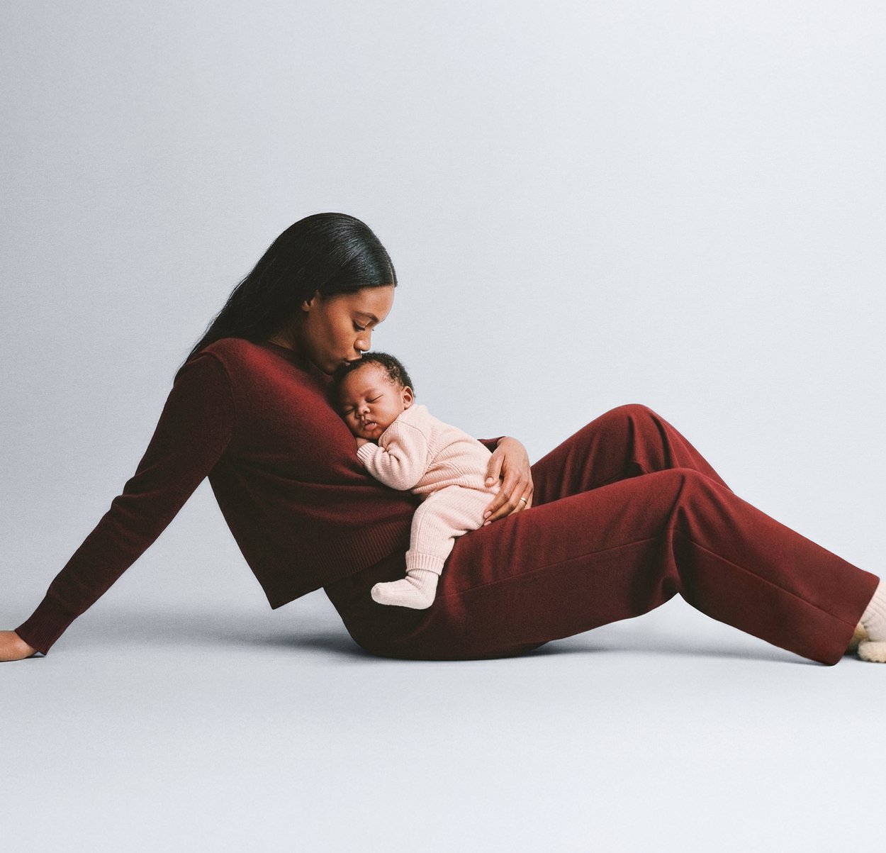 Pulover și pantaloni tricotati bordo purtate de o femeie, cu un bebeluș îmbrăcat într-o ținută roz deschis care doarme în poala ei. Fundal simplu.