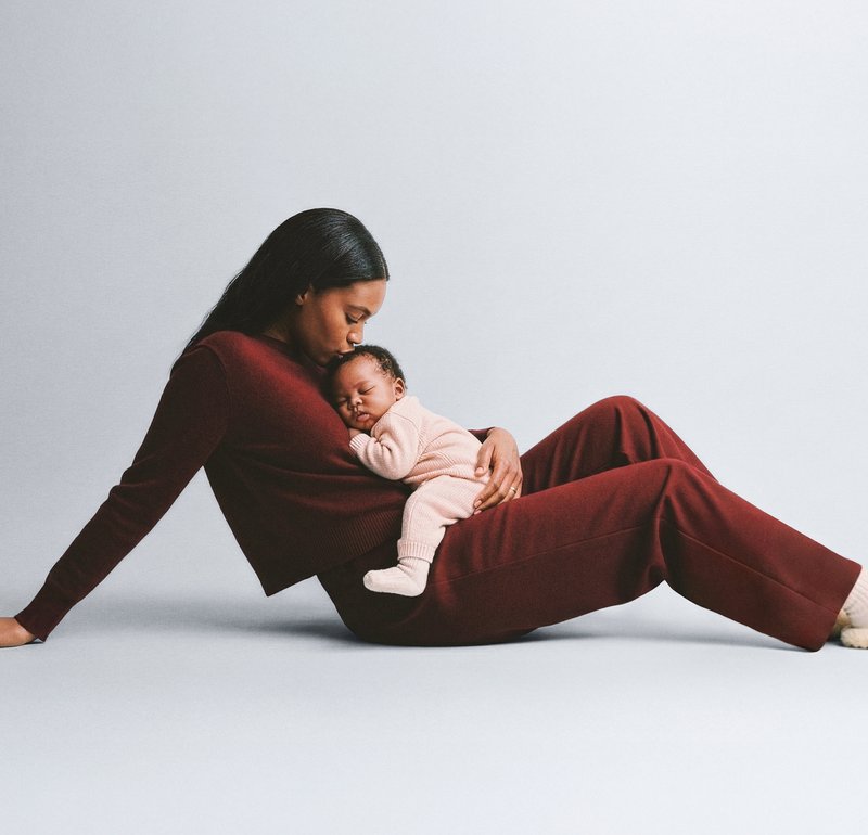 Kõrge kaelusega bordoopunane kootud kampsun ja püksid, mida kannab naine, kes hoiab süles vastsündinut heleroosas riietuses magamas. Lihtne taust.