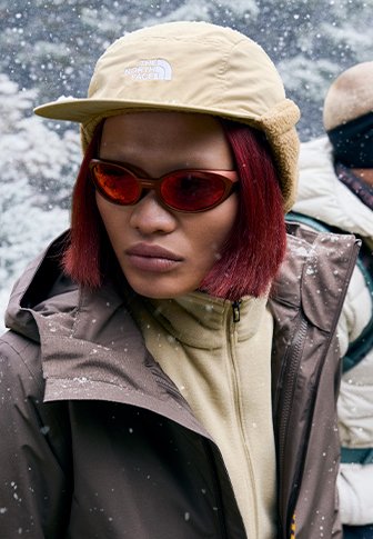 Καφέ αδιάβροχο μπουφάν, μπεζ καπέλο με λογότυπο, κόκκινα ηλιογυαλιά και ανοιχτό μπεζ φλις με φερμουάρ; νιφάδες χιονιού πέφτουν στο φόντο.