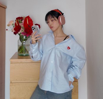 Młoda osoba w za dużej niebieskiej koszuli i różowo-złotych słuchawkach, patrząca na smartfon, stojąca obok drewnianej komody z wazonem na kwiaty.