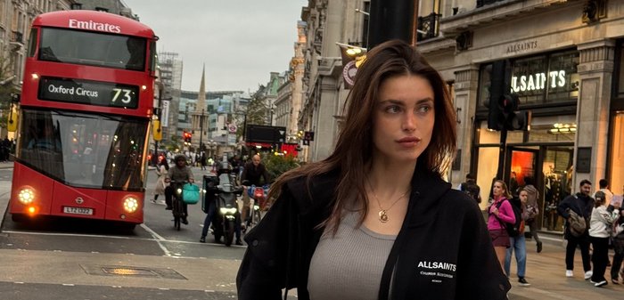 Giacca nera con zip di AllSaints con logo, indossata sopra un top a costine grigio chiaro. Scena urbana con un autobus a due piani rosso sullo sfondo.