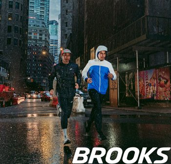 Twee mannelijke hardlopers in een regenachtige stedelijke omgeving. De één draagt een zwart shirt met lange mouwen en de ander een blauw-witte capuchonjack. Het asfalt weerspiegelt het licht.