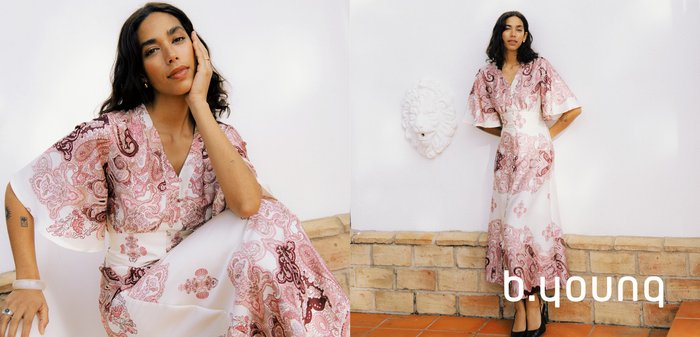 Frau mit langen dunklen Haaren, die ein fließendes rosa-weißes Paisley-Kleid trägt und mit einer Hand ihr Gesicht vor einem schlichten Hintergrund stützt.