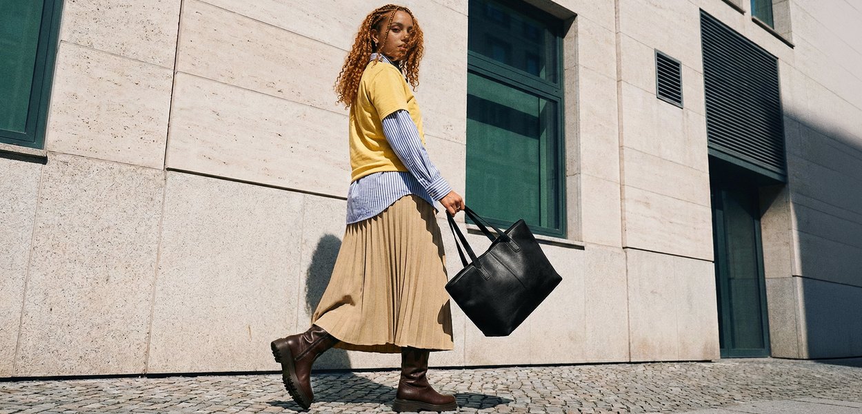 Jeune femme aux cheveux bouclés marchant sur une rue pavée, portant un grand sac fourre-tout noir devant une façade de bâtiment beige.