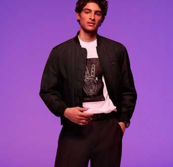 Czarna kurtka bombowca na białym t-shircie z graficznym nadrukiem. Ciemne spodnie i pasek. Fioletowe tło podkreśla kontrast wizualny.