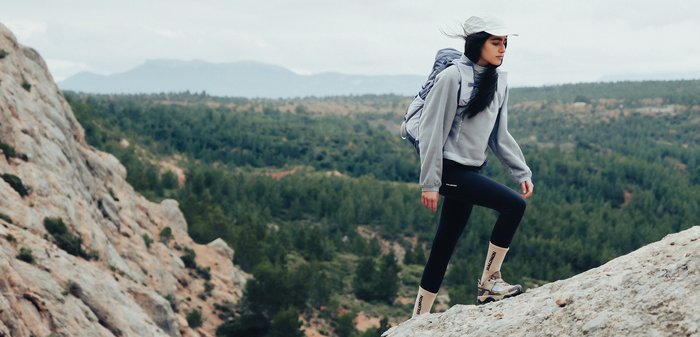 Kvinde iført kasket og rygsæk vandrer op ad en stejl, stenrig bakke med skovklædte bakker i baggrunden.